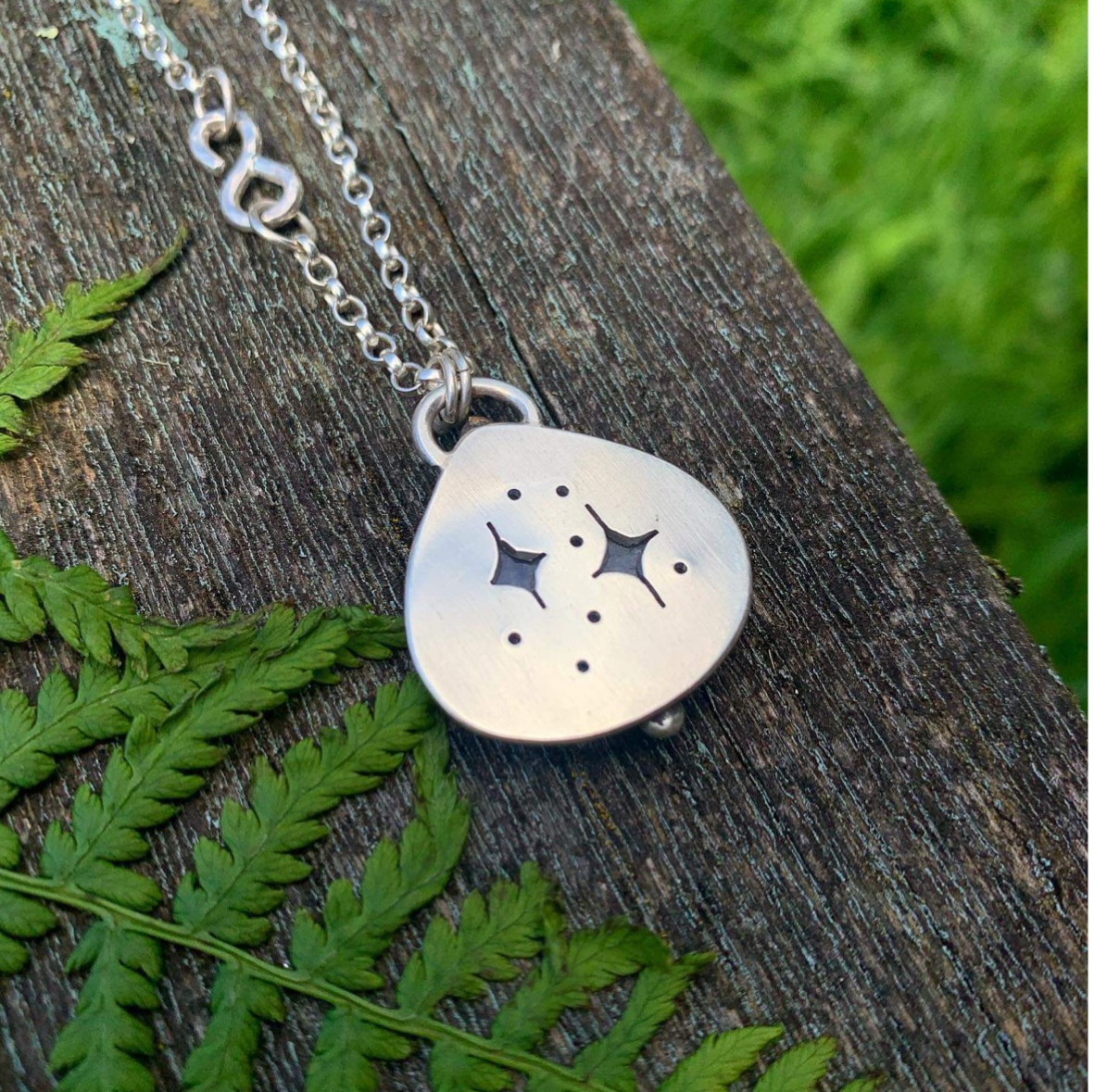 An image of a silver necklace on a piece of oak, with several ferns laid in the background for colour. The pendant of the necklace is a thick teardrop shape with 2 twinkling stars cut out of the center of the pendant revealing the sea glass that is set in the necklace on the reverse